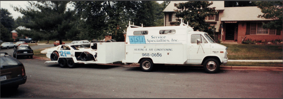 Vintage SSI service van towing a race car on a trailer in a suburban neighborhood