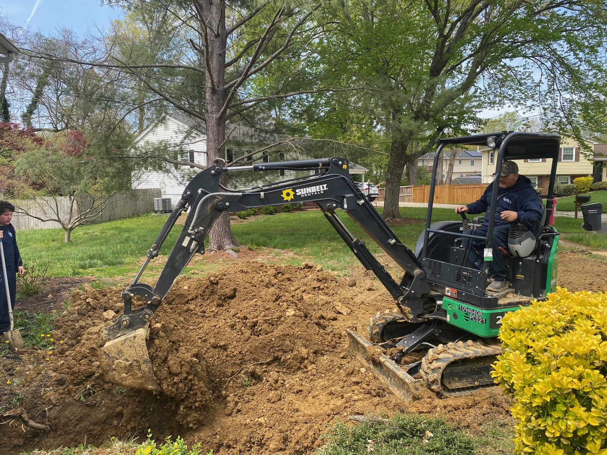 Worker operating a mini excavator to dig soil in a residential yard while another person observes